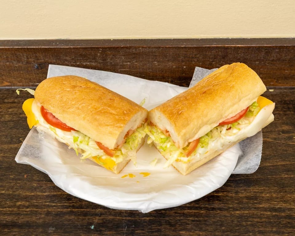 A submarine sandwich, cut in half, with lettuce, tomato, and cheese on a white paper plate.