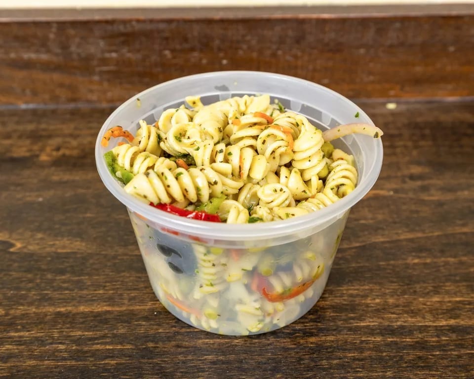 A clear plastic container filled with pasta salad, featuring rotini, vegetables, and herbs.