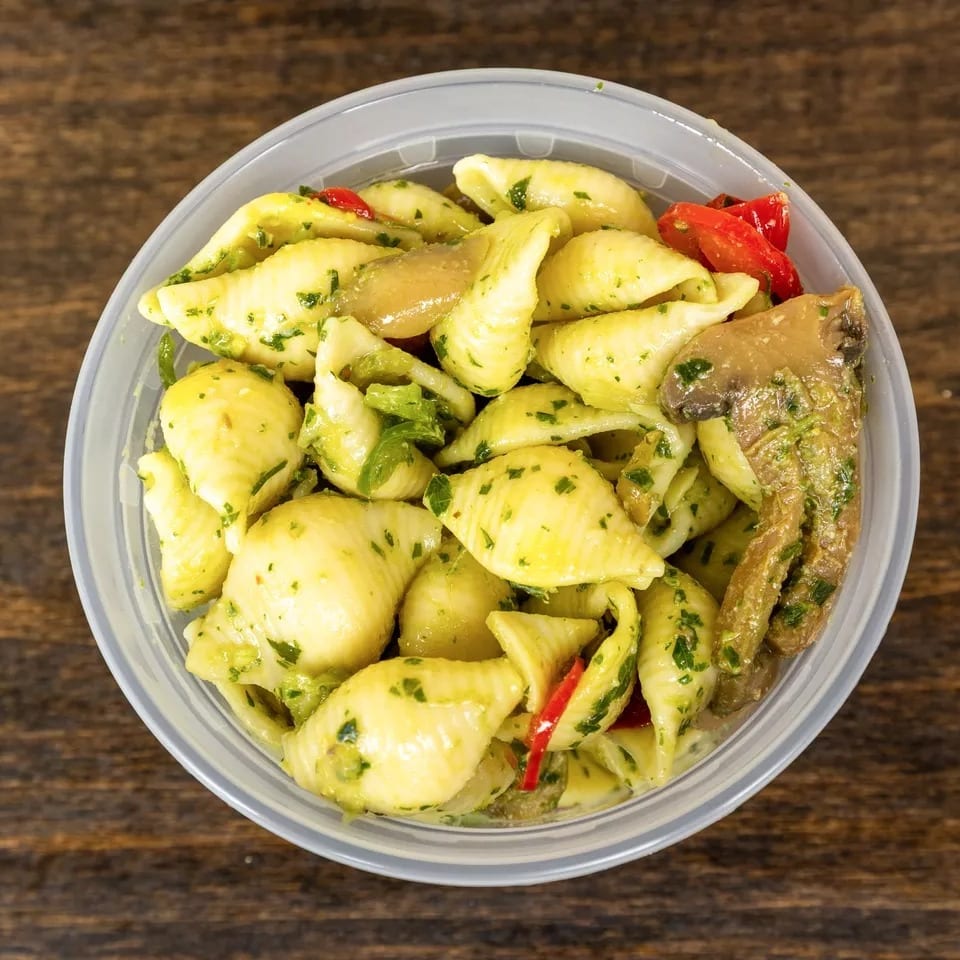 Top-down view of pasta salad with shell pasta, pesto, mushrooms, and red peppers in a plastic container.