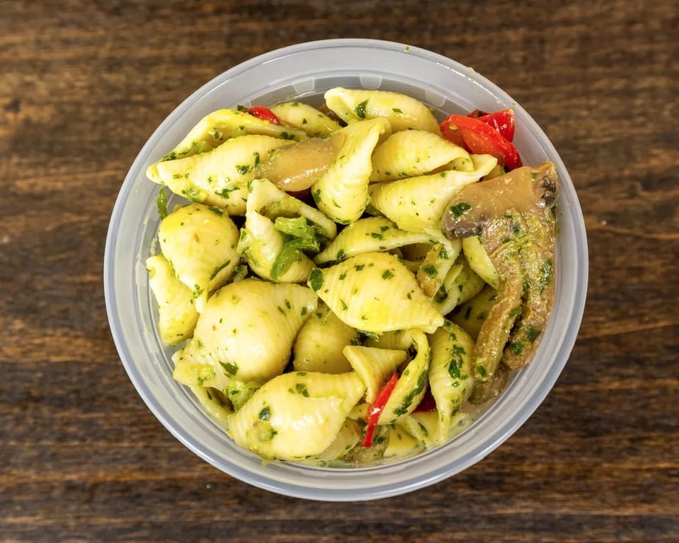 Top-down view of pasta salad with shell pasta, pesto, mushrooms, and red peppers in a plastic container.