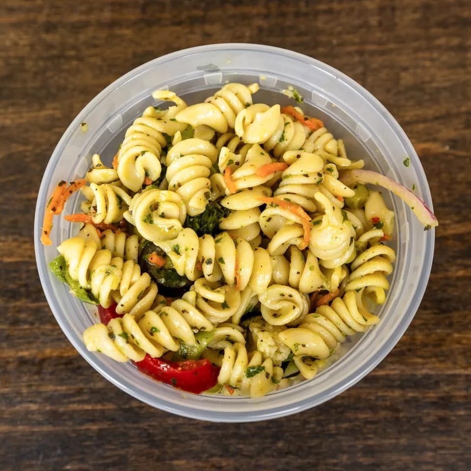 Top-down view of a clear plastic container filled with pasta salad on a wooden table.