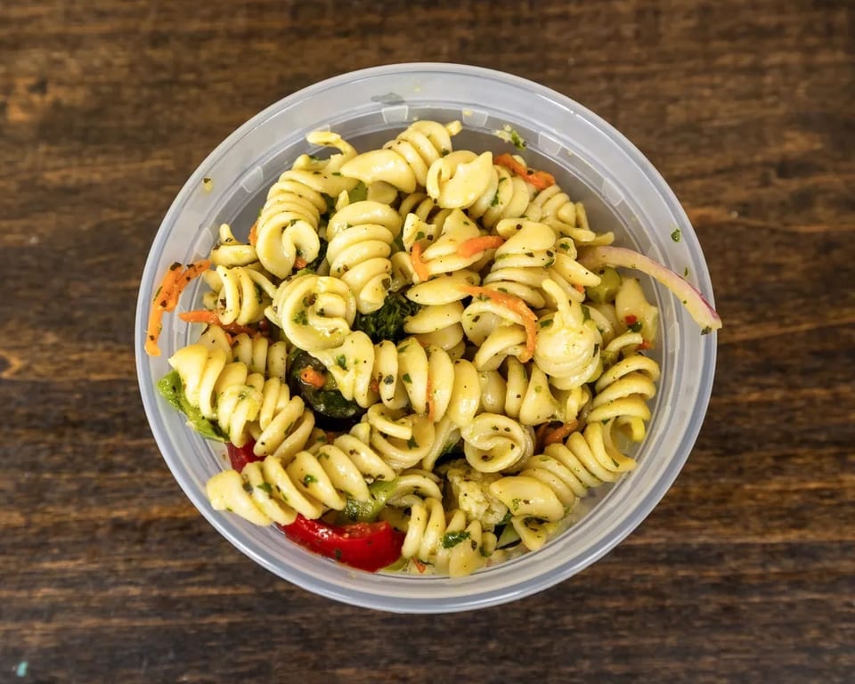Top-down view of a clear plastic container filled with pasta salad on a wooden table.