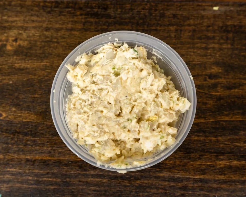 Creamy potato salad with visible chunks of potato and green specks in a clear plastic container.